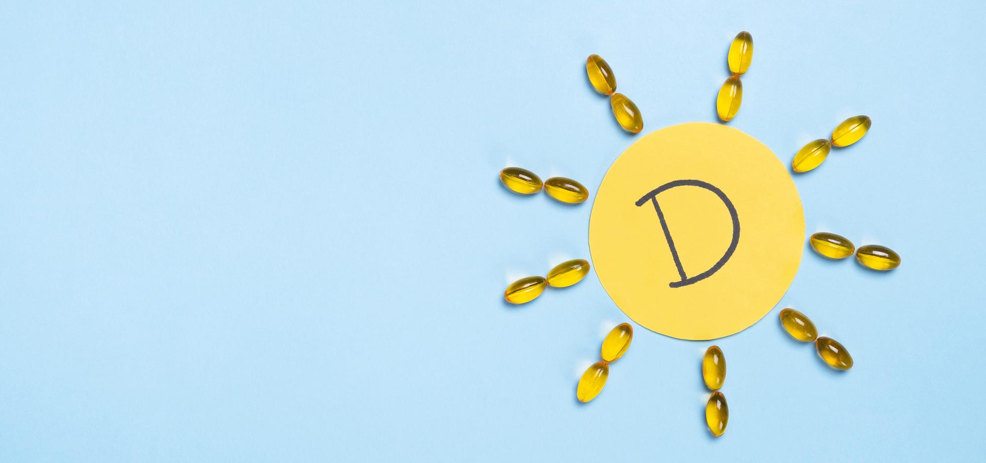 D-vitamin i gullfargede kapsler arrangert som solstråler rundt en gul sirkel med bokstaven D, på en lyseblå bakgrunn som symboliserer solens rolle i D-vitaminproduksjonen.